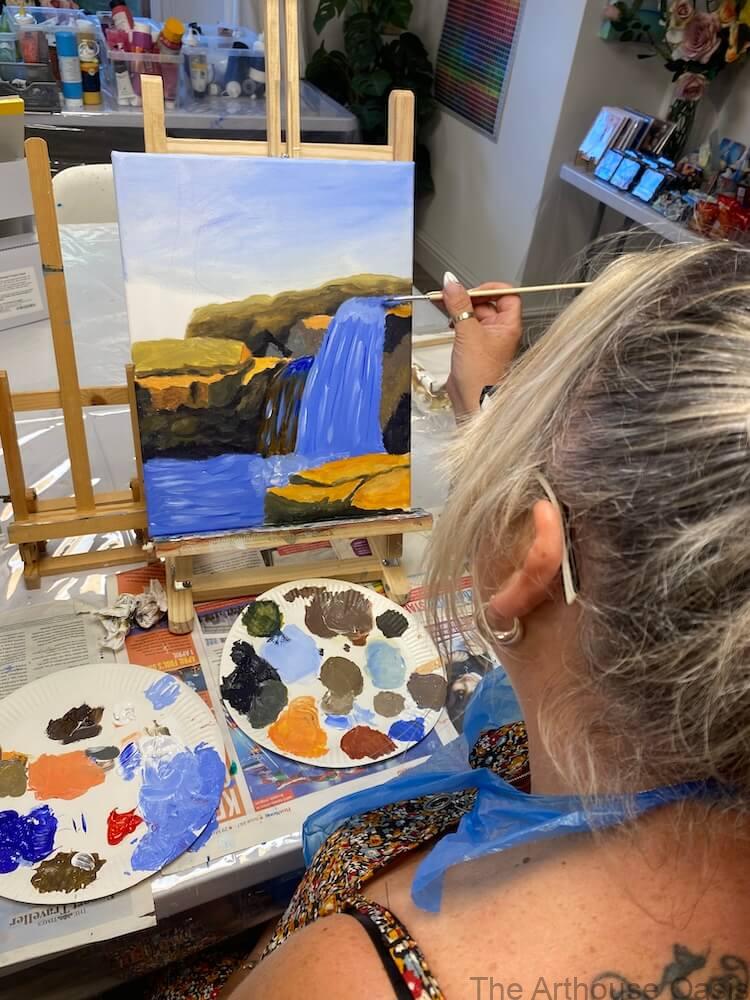 Alt Text:
Student painting a waterfall and rocky landscape in acrylics during an art class.