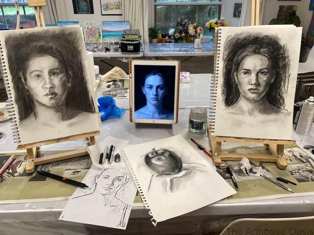 Two charcoal portrait drawings on easels with a tablet reference photo in between