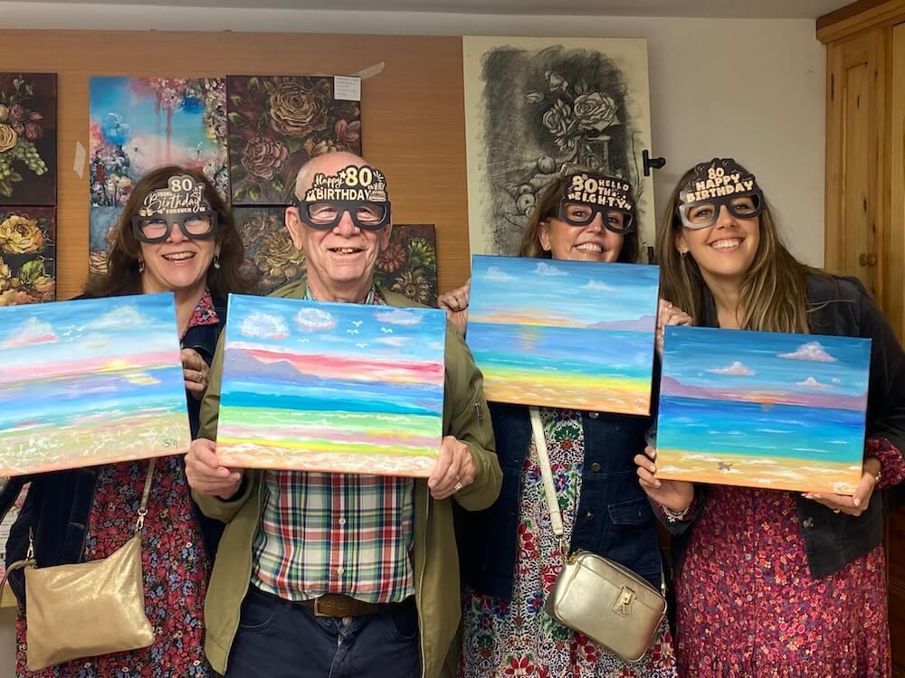 Four smiling participants wearing birthday glasses holding up ocean paintings.
