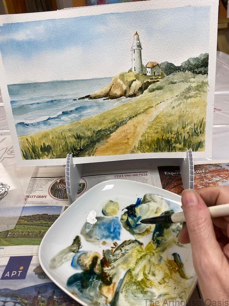 Hand holding a brush over a palette with a coastal watercolour painting of a lighthouse on an easel.