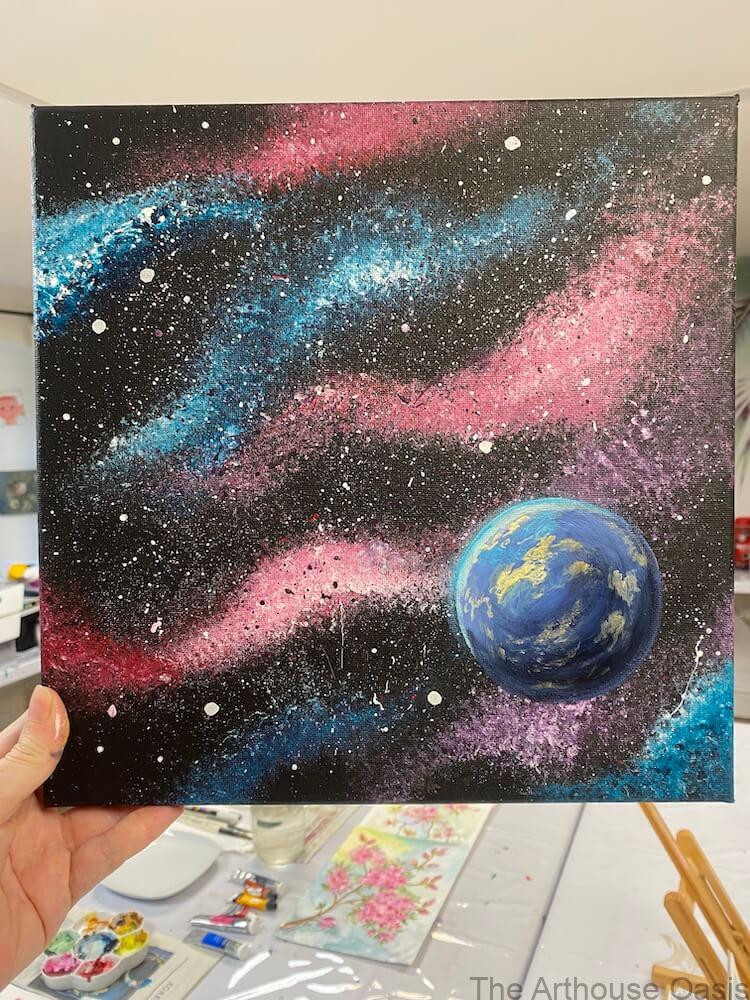 A finished acrylic galaxy painting with pink and blue nebulas and a bright blue planet, held up for display.