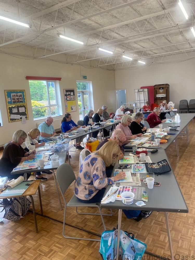 A lively watercolour workshop with participants painting at long tables in a bright, spacious hall.