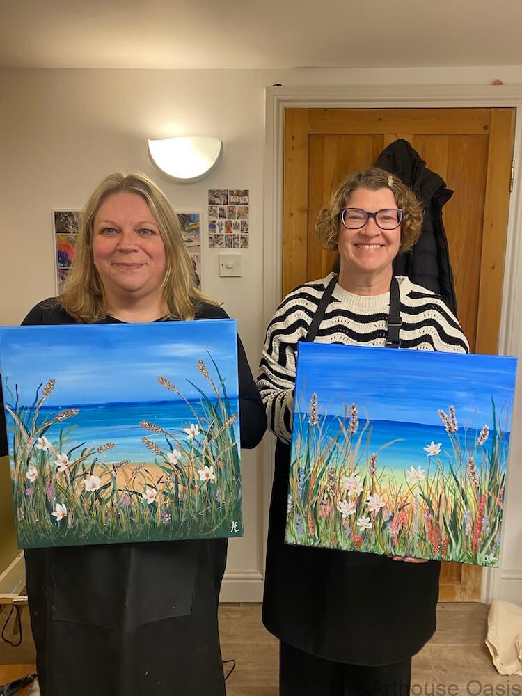 Two finished acrylic coastal paintings held by artists after completing a guided art session.