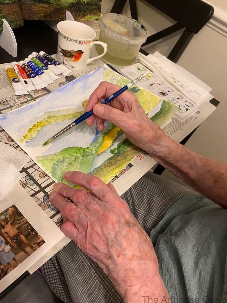 Close-up of hands painting a watercolour landscape with layered washes on paper.