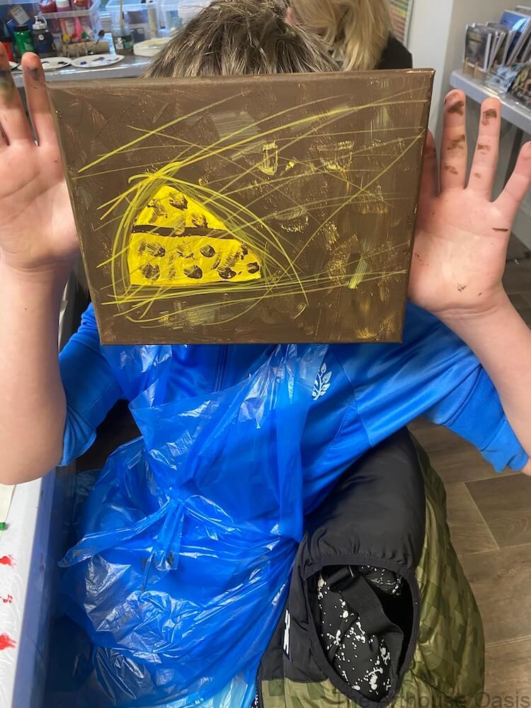 Child holding an acrylic painting of a slice of cheese on a brown background in an art class.