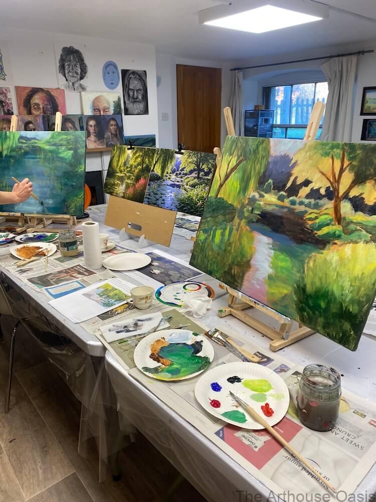 Large-scale impressionist landscape paintings in progress on easels inside an art studio.