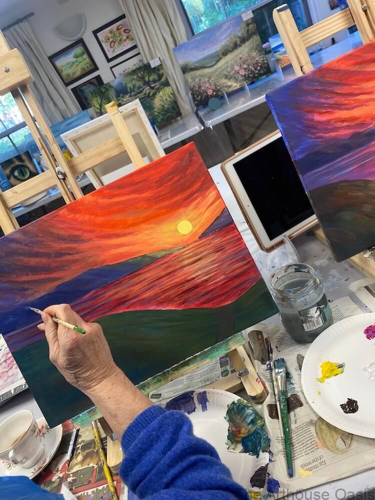 Adult students painting colourful sunset landscapes on canvas during an acrylic art class in Bishop’s Stortford.