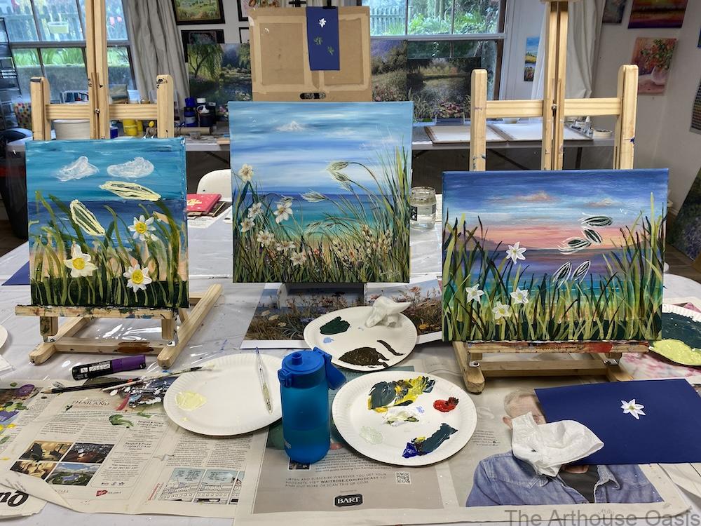Group of coastal sunset acrylic paintings with flowers and grasses on easels in an art studio.